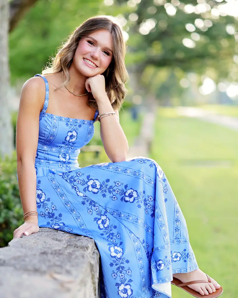 Lily Butler in a blue floral dress, sitting on a stone wall in a park and smiling