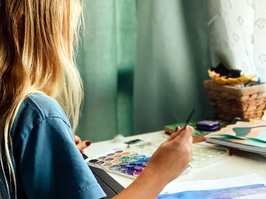 A young student painting with watercolors at a table by a window